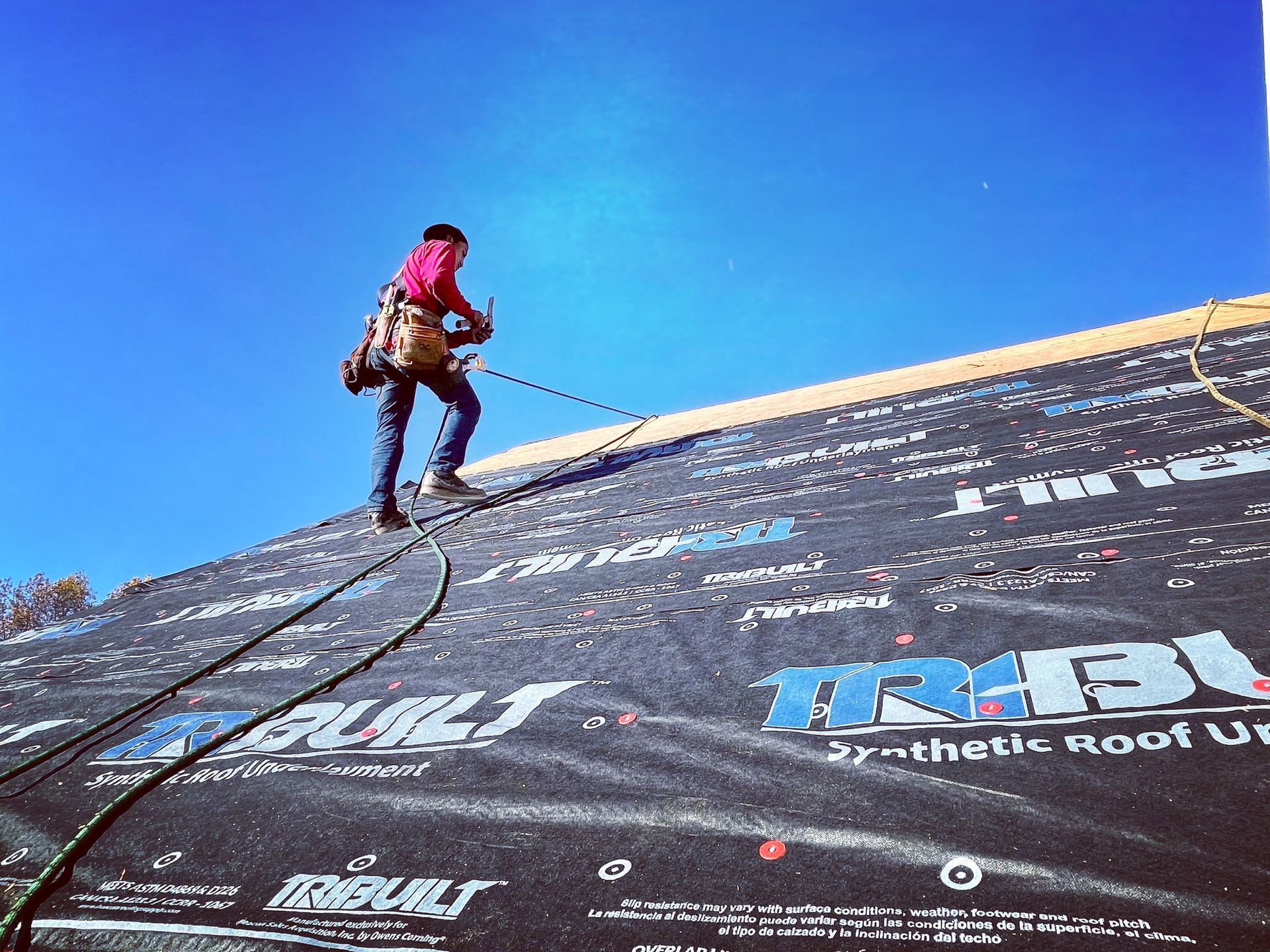 Worker on a steep rooftop under a blue sky, securing roofing material with a harness.
