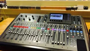 Audio mixing console with many sliders, knobs, and a small display in a studio setting