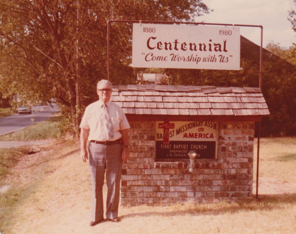 Man standing beside a brick roadside sign reading “Centennial” in a sunlit rural setting