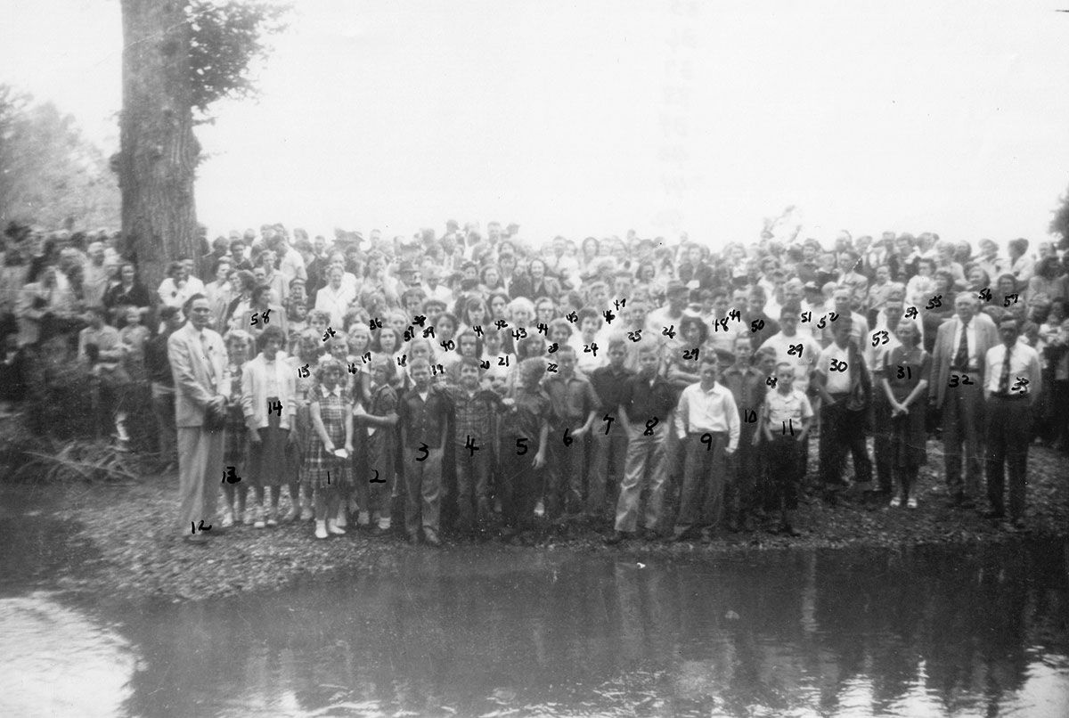 Large crowd standing by a riverbank near trees, reflected in the calm water