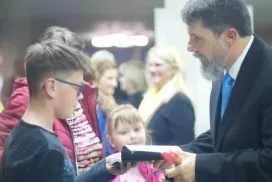 Man in suit hands a certificate to a boy in glasses during a ceremony, with people blurred in the background
