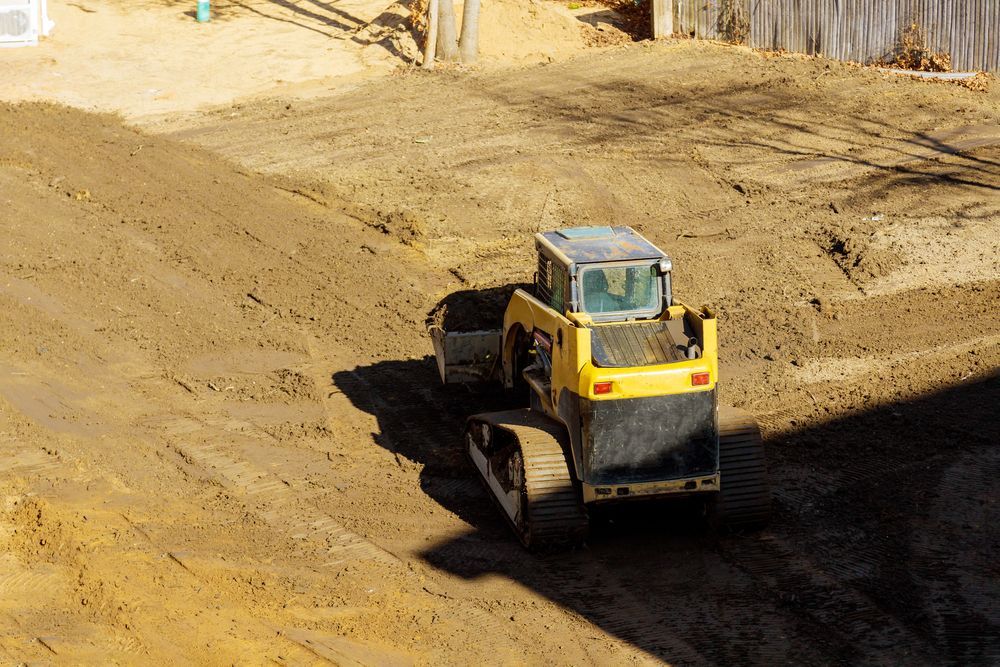 A yellow tracked skid steer loader works on a dirt construction site, clearing and grading uneven ground.