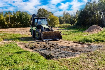 A green tractor with a front bucket sits on a gravel patch in a grassy field near a forest on a bright day.