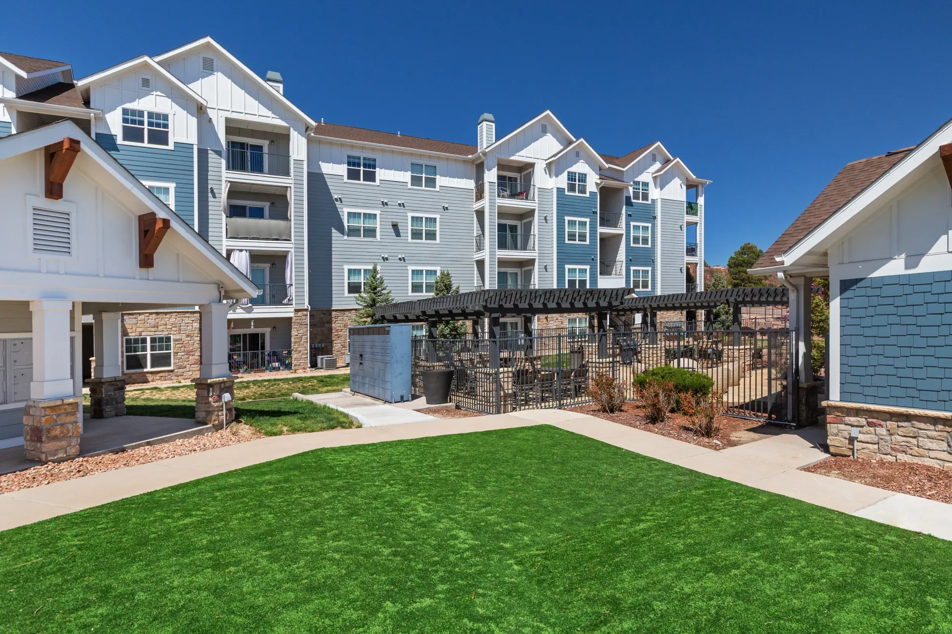 Exterior view of an apartment complex with a fenced outdoor seating area and green lawn.