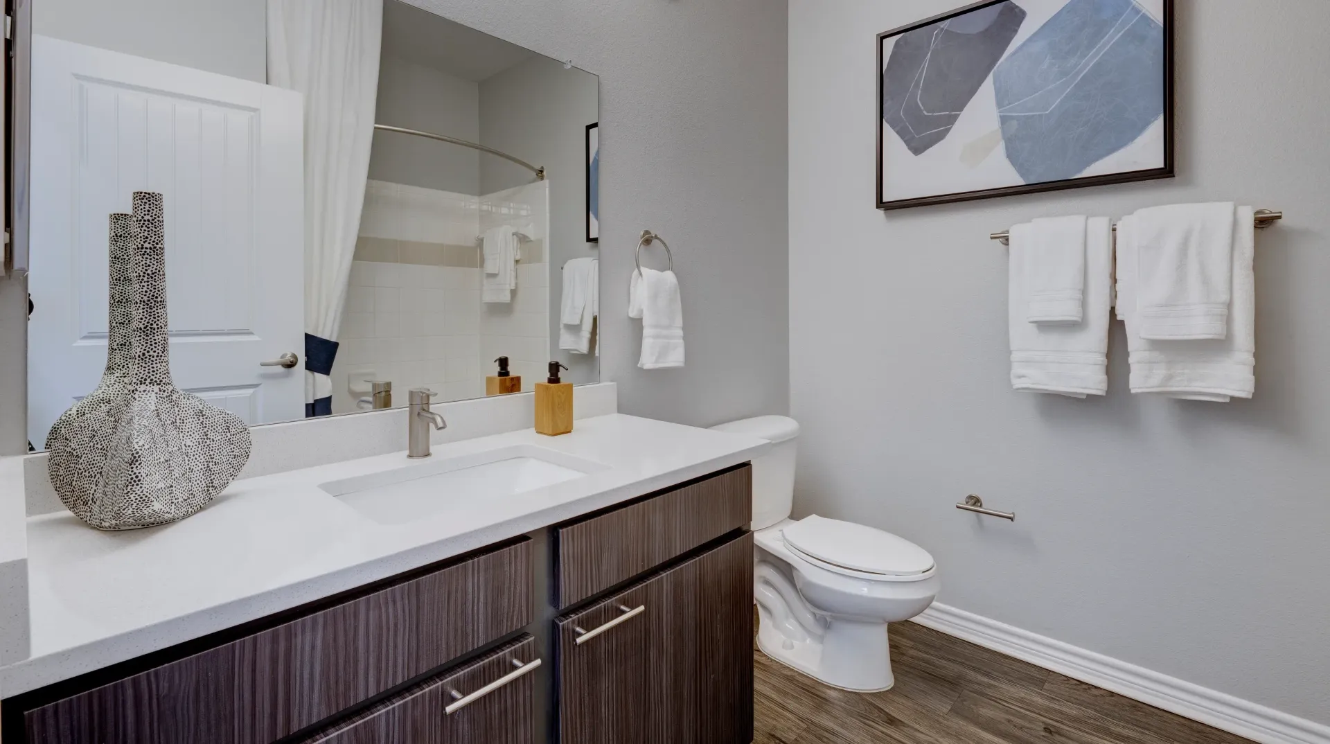 Modern bathroom vanity with white countertop, dark wood cabinets, and a silver faucet. Toilet and artwork visible.