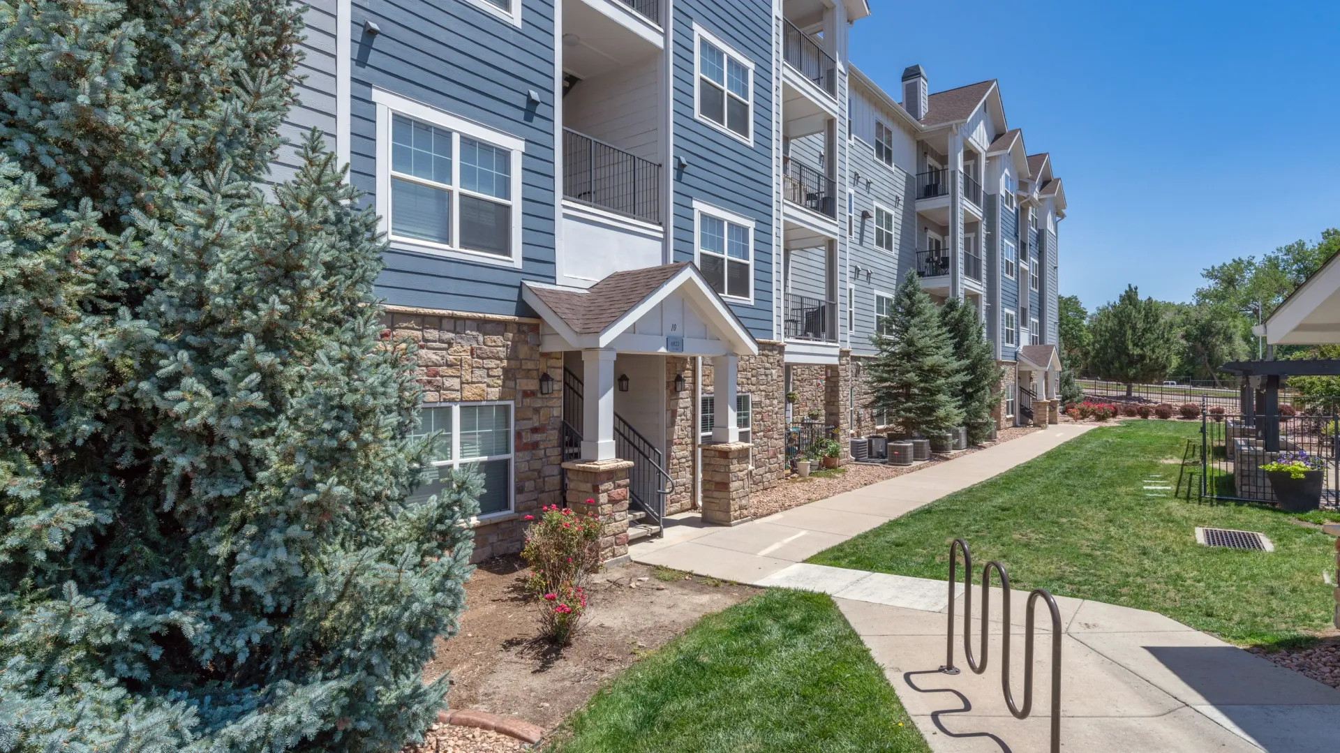 Exterior view of apartment building entrance with blue siding and stone accents, surrounded by trees and landscaping.