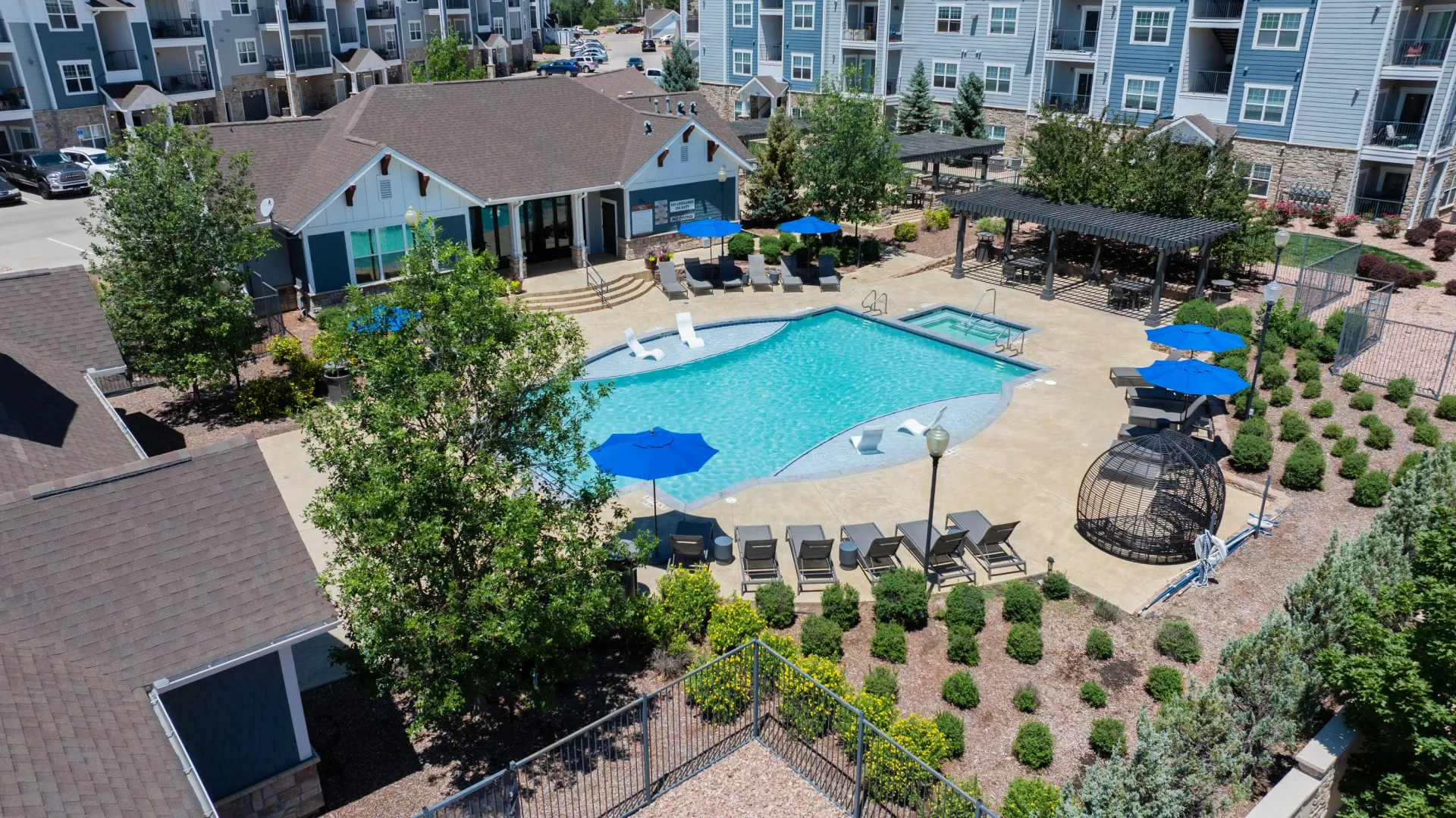 Aerial view of a resort-style swimming pool with lounge chairs and umbrellas, adjacent to apartment buildings.