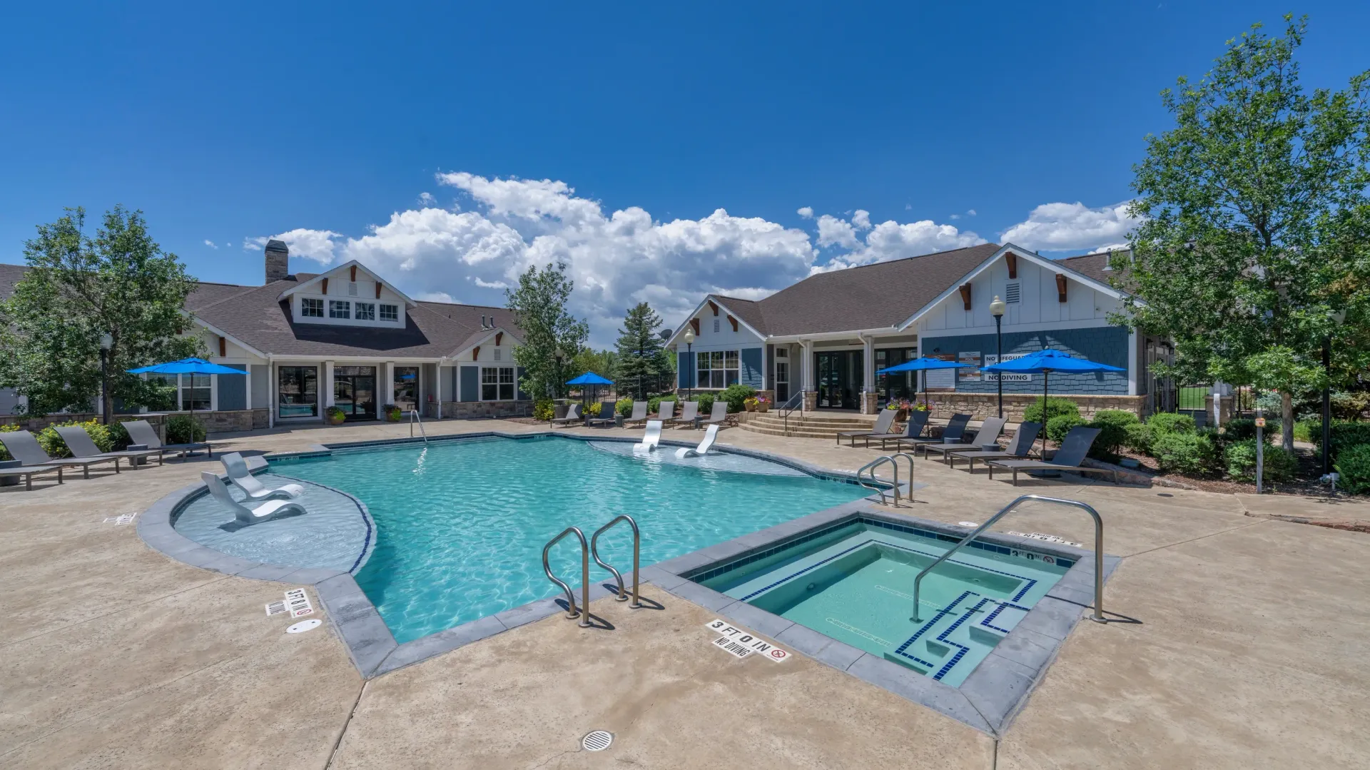 Outdoor swimming pool and hot tub area with lounge chairs and umbrellas, adjacent to apartment buildings.