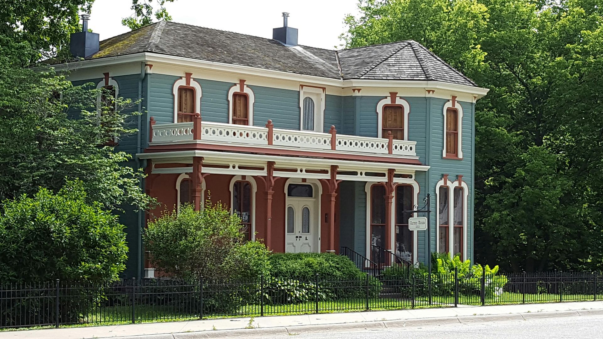 Historic two-story brick house with white trim and red columns behind trees and a fence