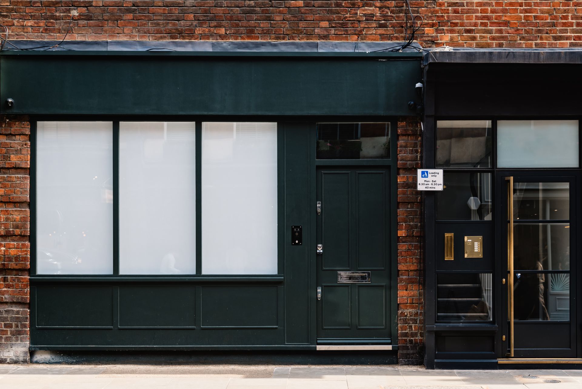 Dark green storefront with white-covered windows and door next to a black doorway, all against a brick wall. Dark green storefront with white-covered windows and door next to a black doorway, all against a brick wall.