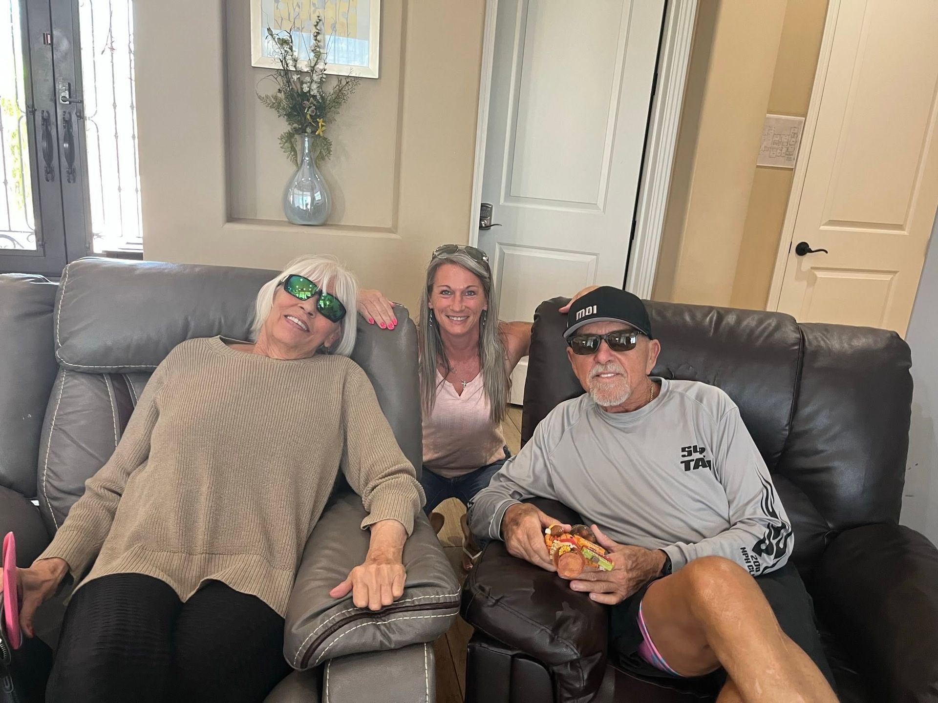 Three people relax on a couch, smiling in a bright living room.