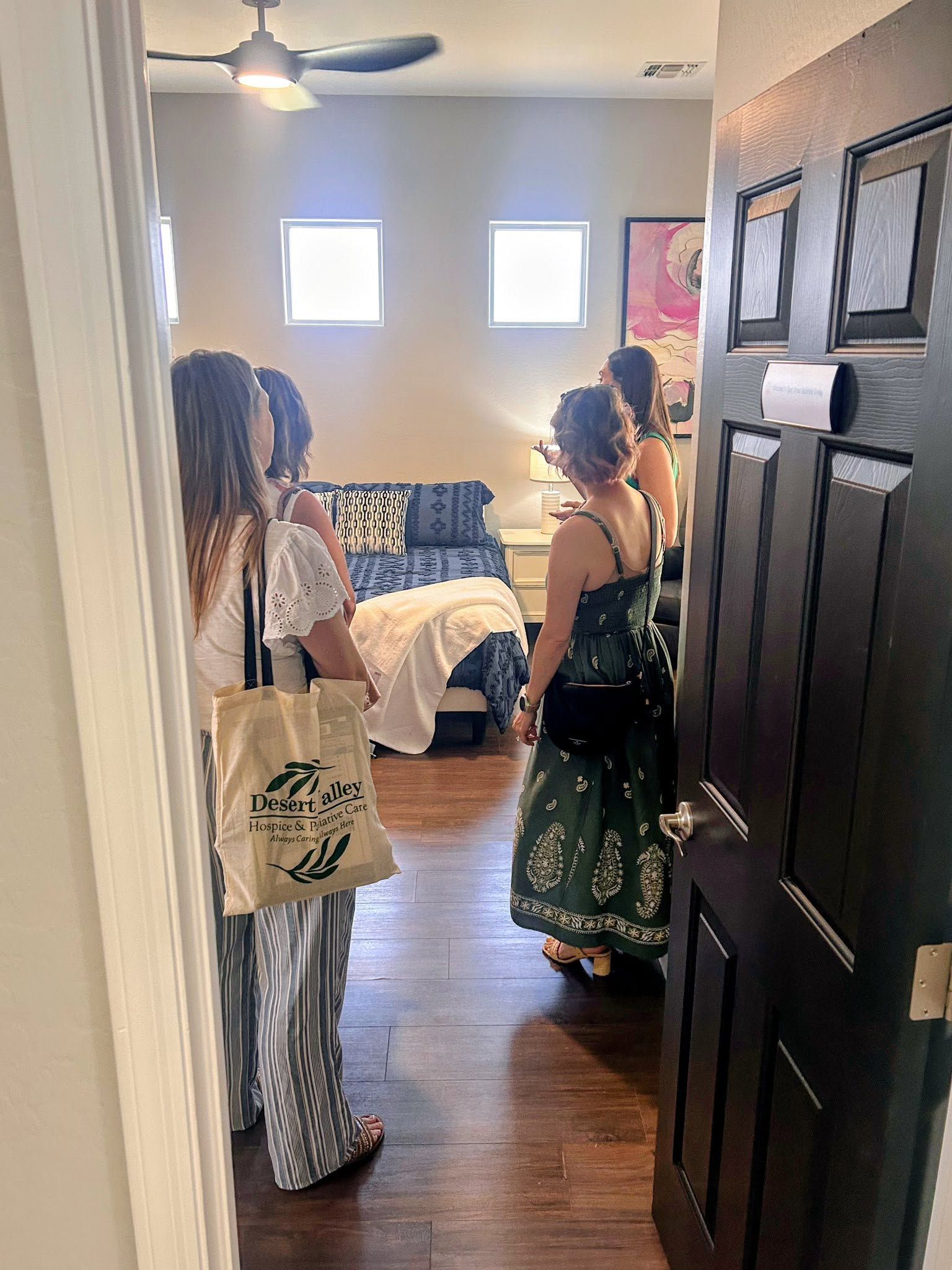 Two women standing in a bedroom doorway, looking toward a bed with a patchwork quilt.