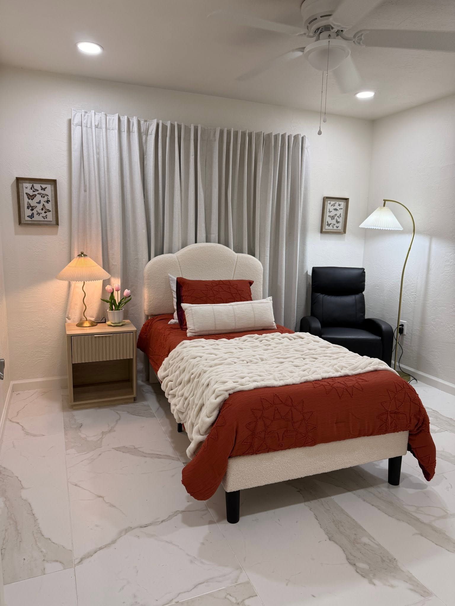 Cozy bedroom with white tufted bed, red bedding, lamp, chair, and curtained window on tiled floor