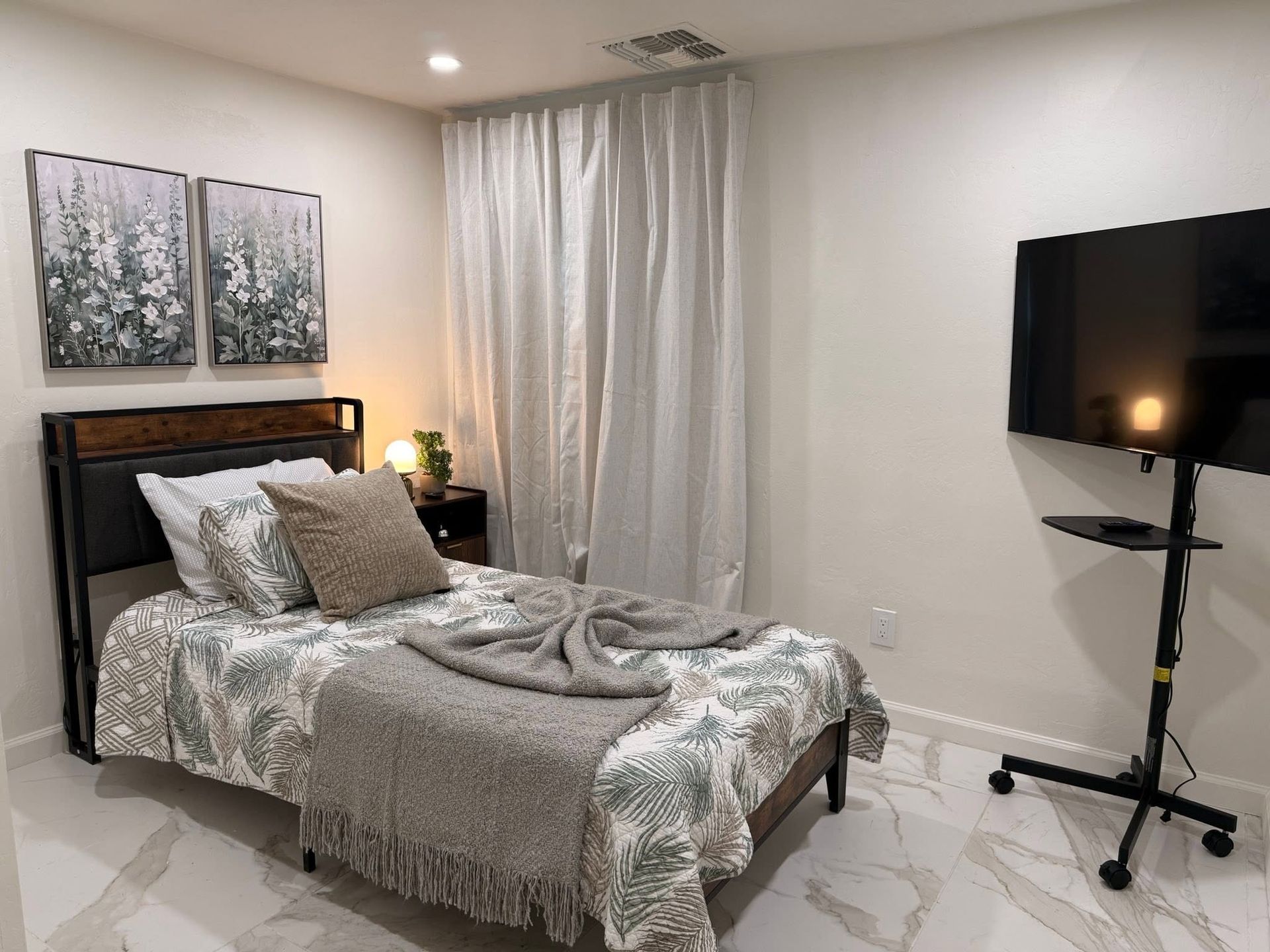 Small bedroom with a bed, bedside lamp, wall art, curtains, and a TV on a stand.