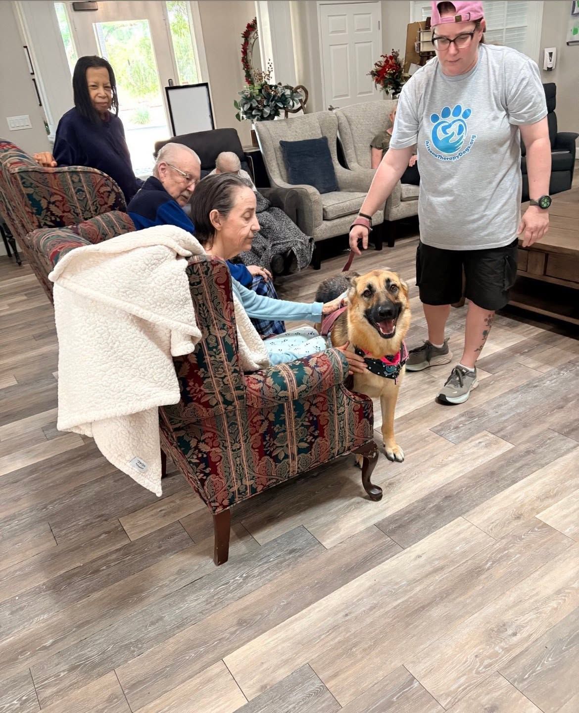 Woman in a chair petting a happy dog in a living room, with two people nearby.