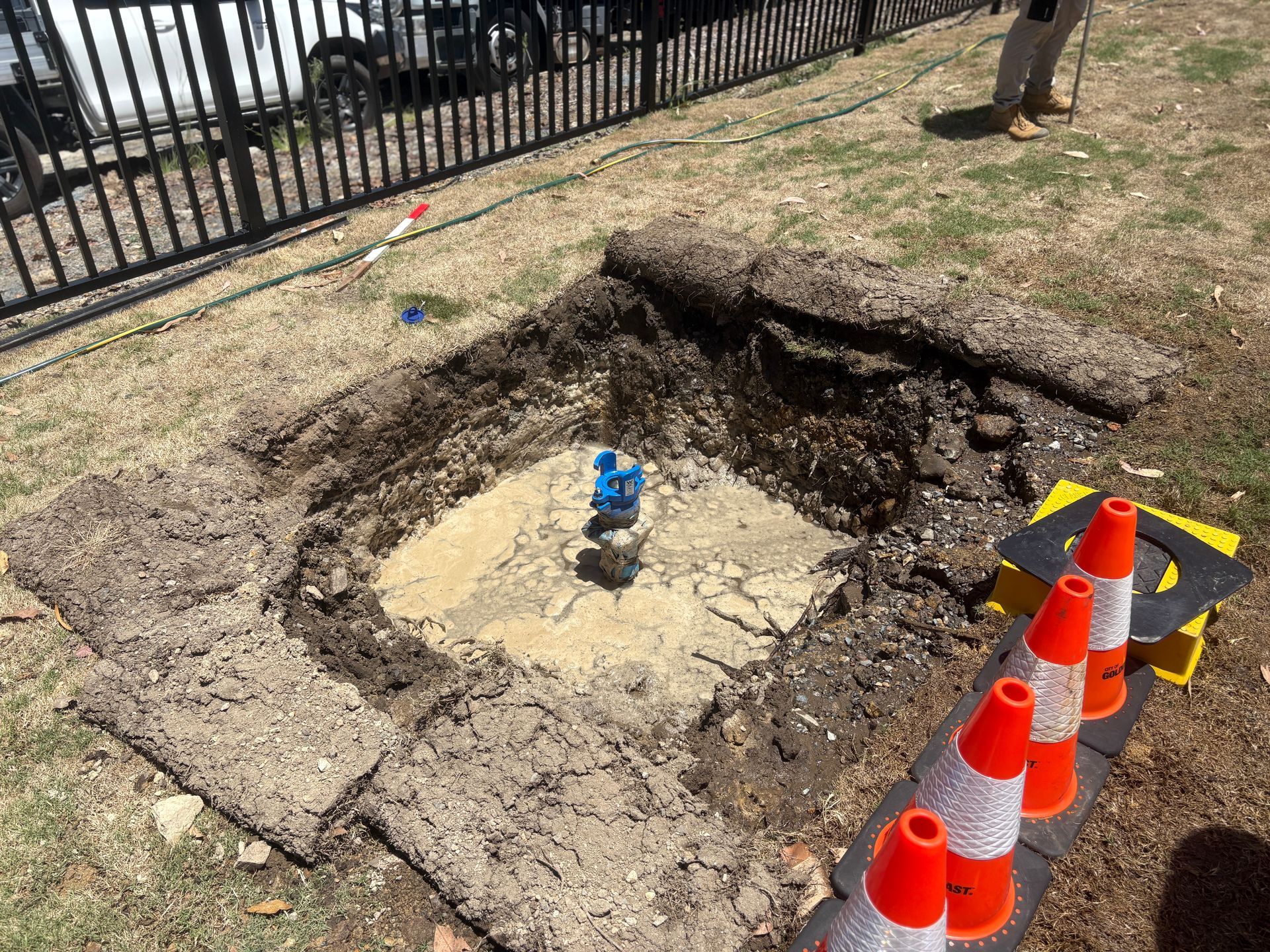 A square excavation in a grassy yard featuring a central pipe fitting, marked by a row of orange traffic cones nearby.