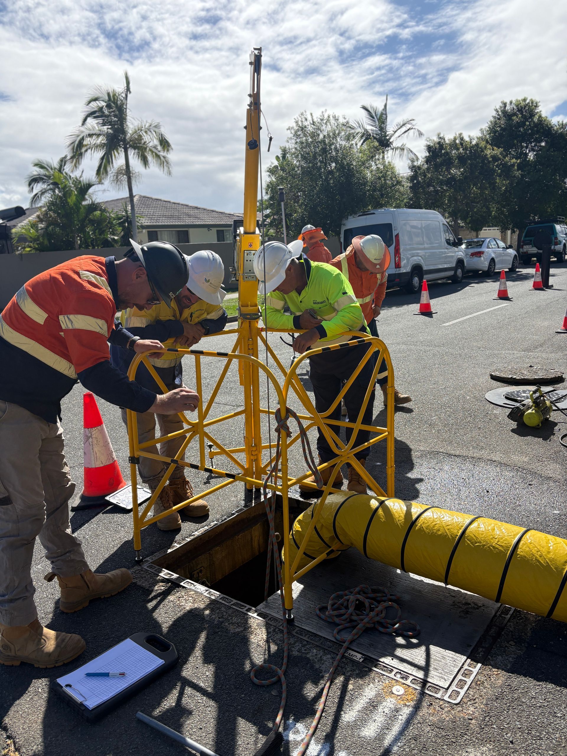 Construction workers in high-visibility gear set up a tripod safety device over an open manhole on a sunny street.