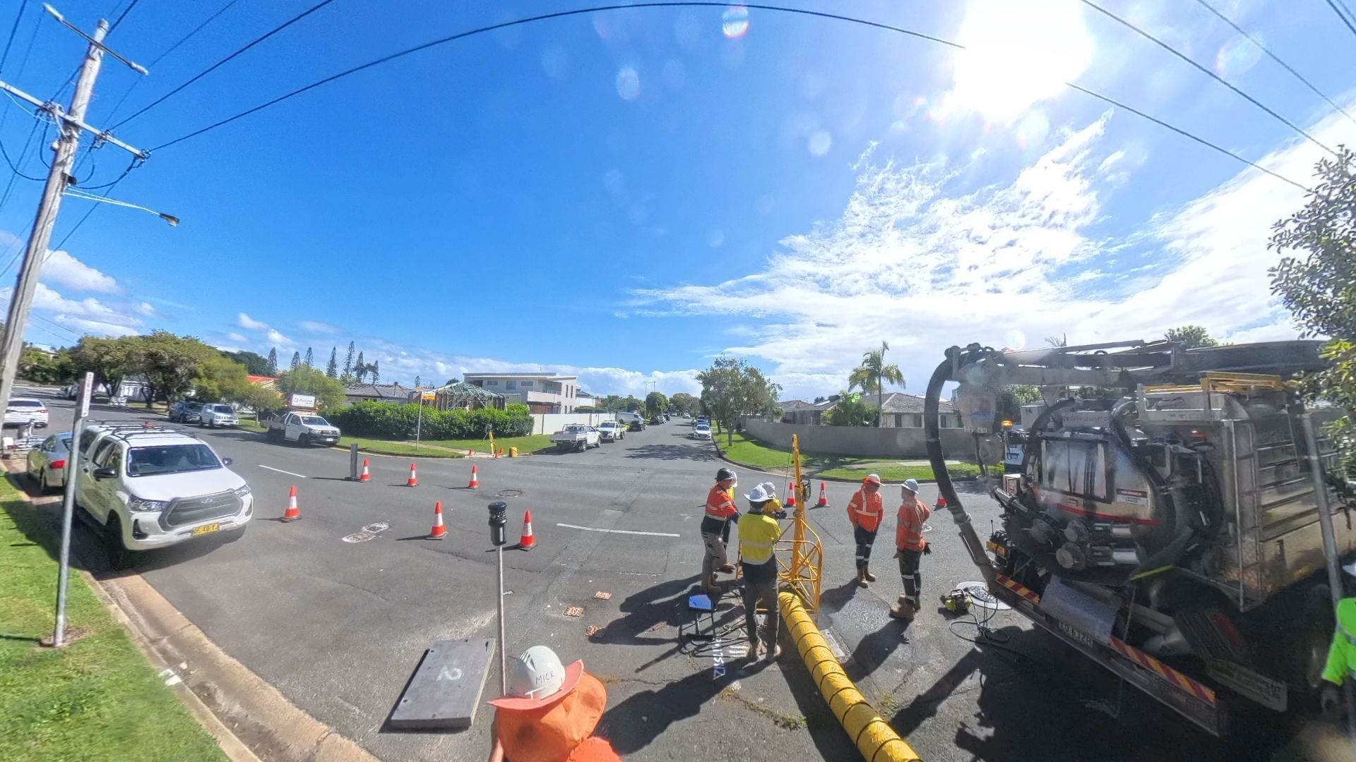 Utility workers in high-visibility vests work near a directional drill machine on a suburban street under a sunny sky.