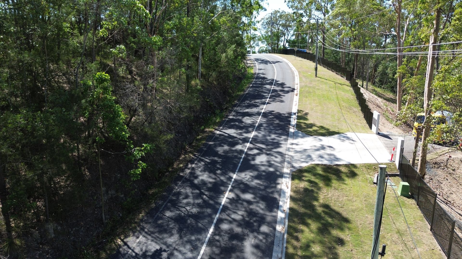 A paved road with a white center line curves through a forest, bordered by grass and a construction fence on the right.