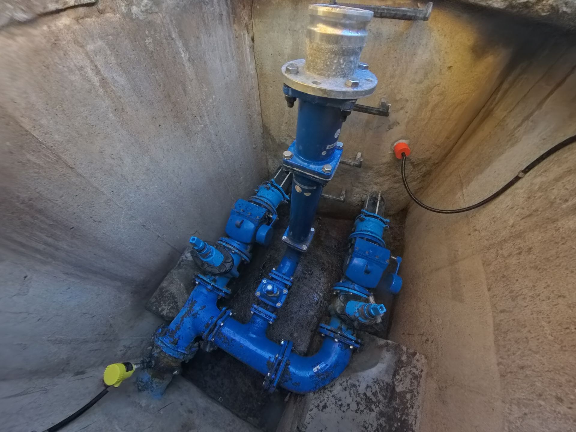 Sewer piping and valves in a concrete pit, with a yellow cap and a clear vertical pipe at the top.