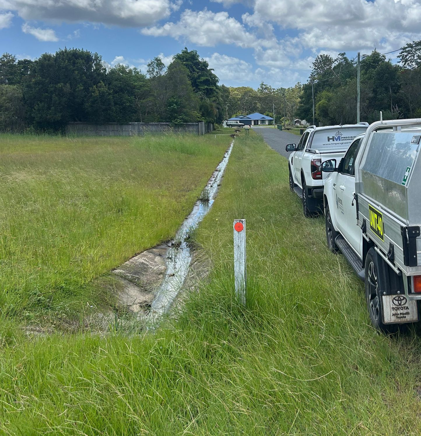 Two white utility trucks parked on a grass verge beside a drainage ditch on a sunny, rural roadside.