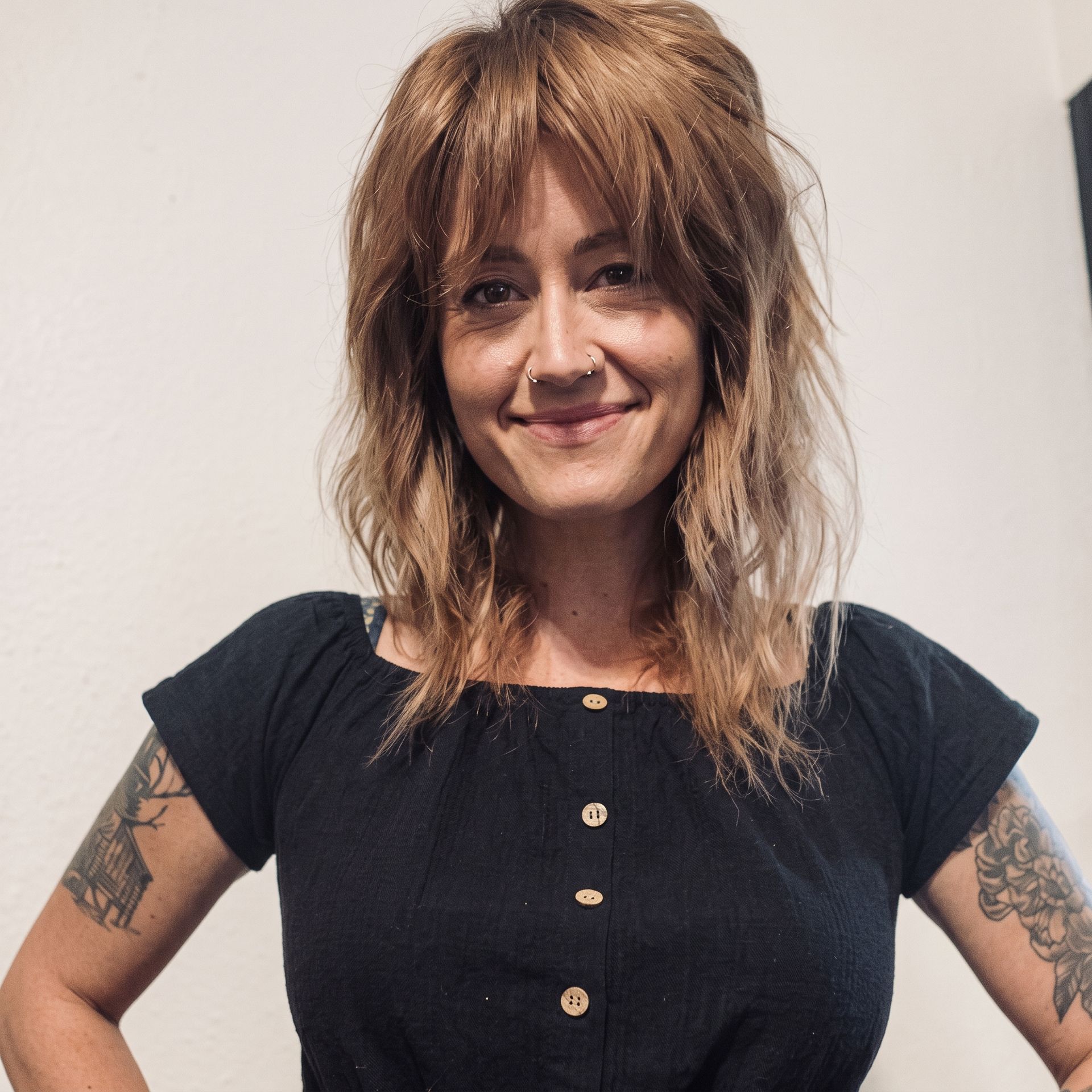 Woman with shoulder-length auburn hair and tattoos, wearing a black top, smiling against a plain wall