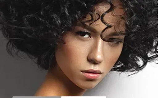 Close-up portrait of a person with curly dark hair and a neutral expression against a gray background