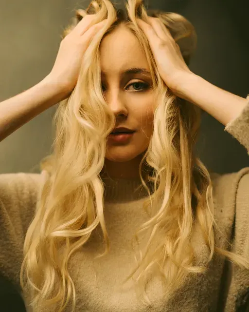 Blonde woman with hands on her head, wearing a beige sweater against a muted background.