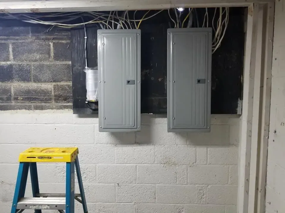 Two gray electrical panels mounted on a cinder block wall, with wires above. A yellow and blue step stool is in the foreground.