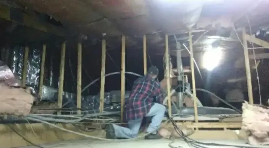 Person working on electrical wiring in an attic, kneeled down. Dark interior with insulation and exposed beams.