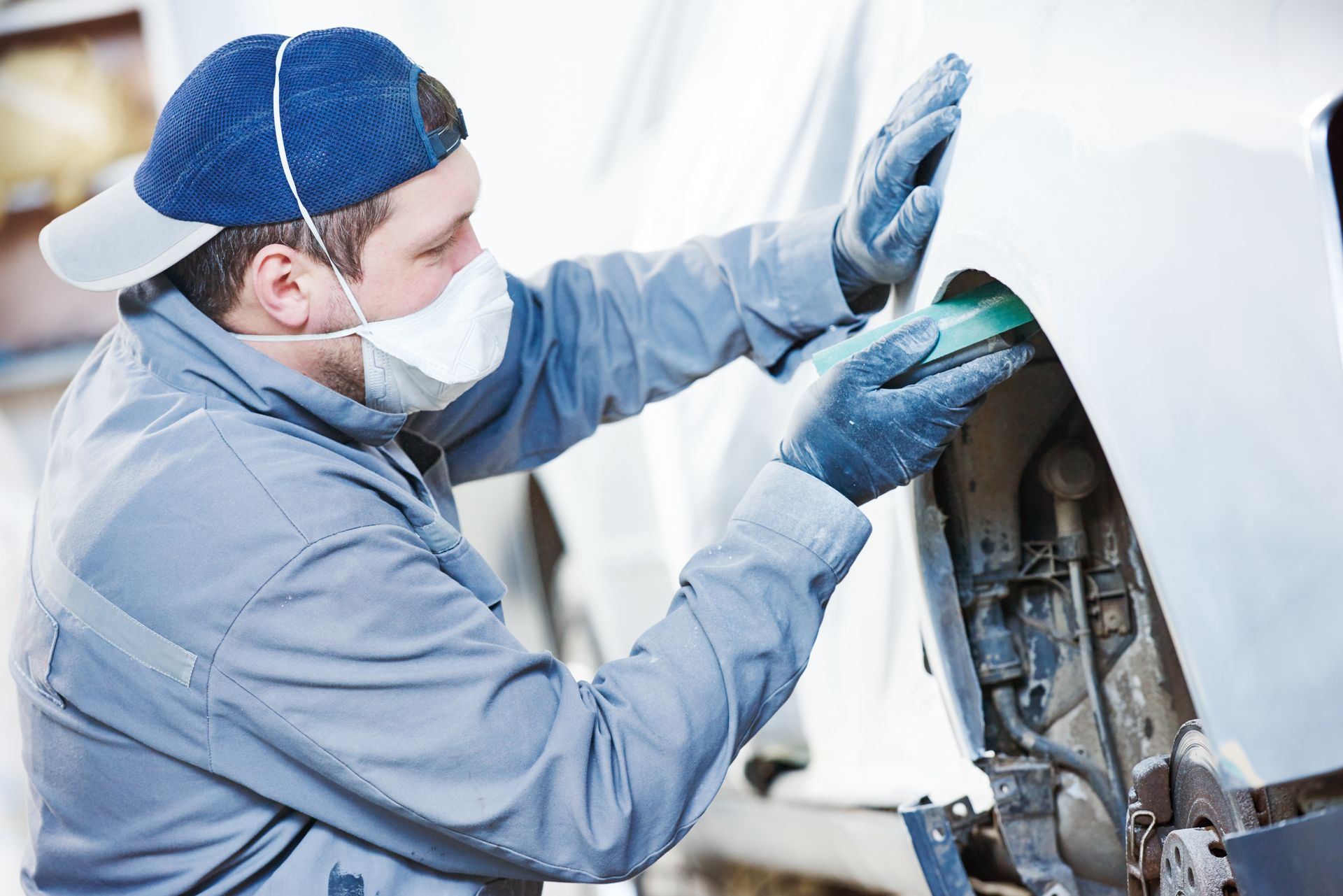 A technician wearing a face mask and gloves sands the body of a car in a repair shop.