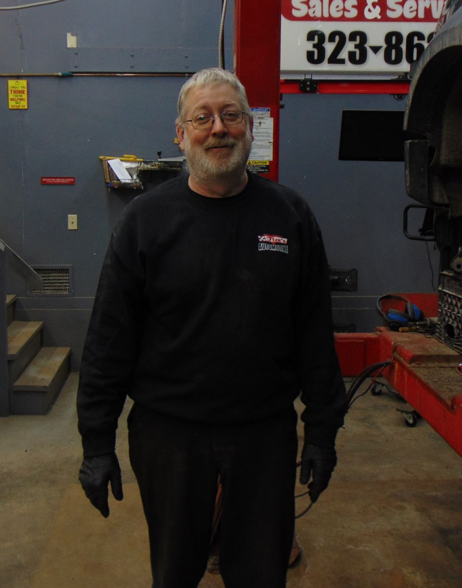 A person in a black sweatshirt and gloves smiles in a vehicle maintenance shop with equipment and a sign in the background.