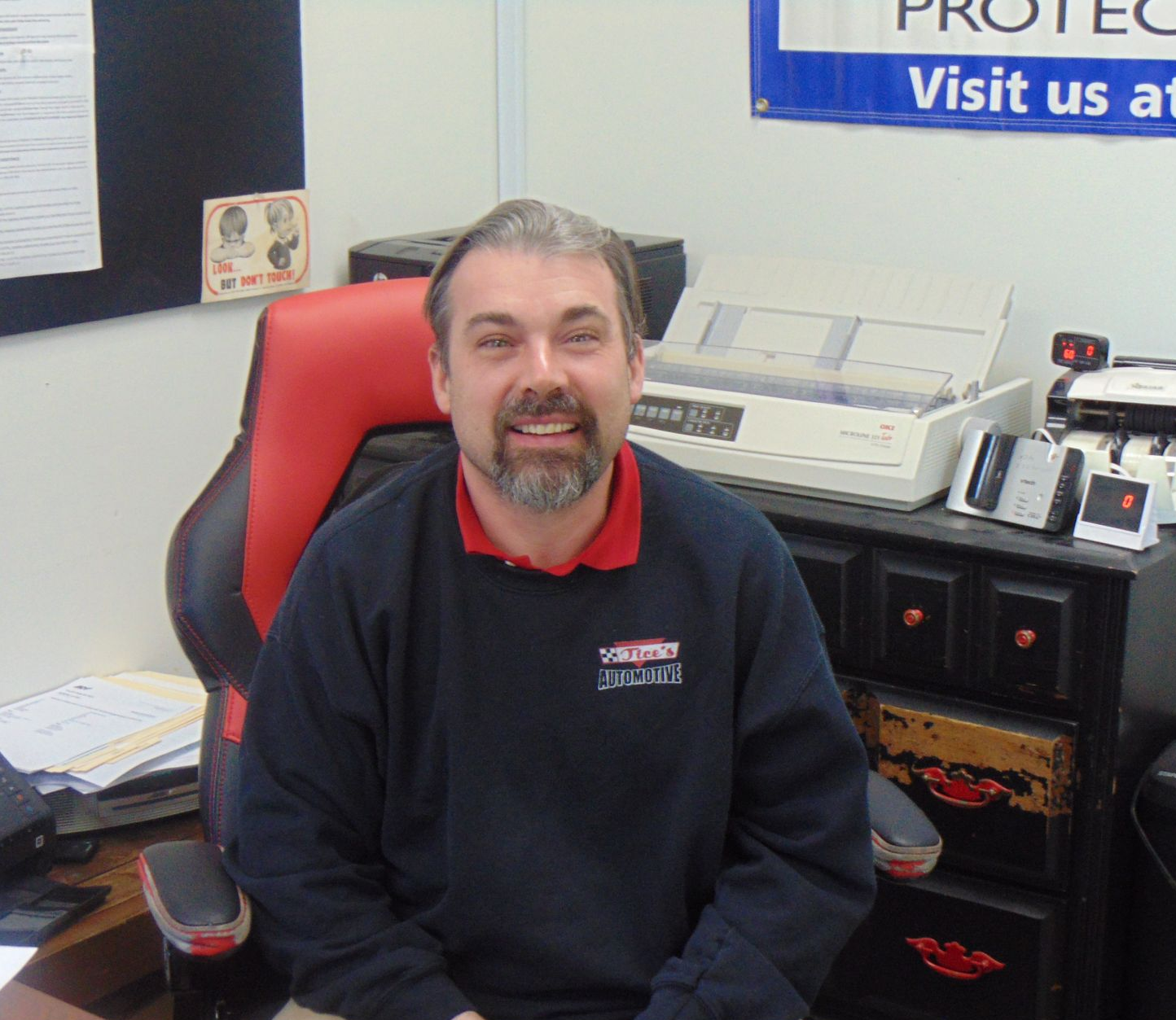 A person with a beard, wearing a dark sweatshirt, smiling while seated in a red and black chair in an office.