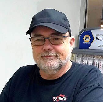 A person wearing a dark cap, glasses, and a logo t-shirt, standing in front of a NAPA auto parts display.
