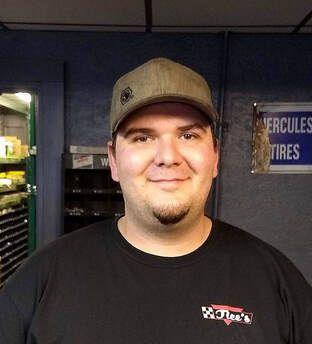 A man in a grey baseball cap and a black t-shirt with a logo stands in front of shelving and a Hercules Tires sign.