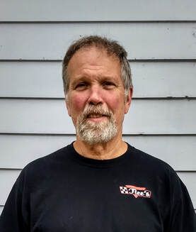 A person with a beard and short hair wearing a black t-shirt, posed in front of light gray horizontal siding.