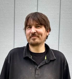 A person with brown hair and a beard, wearing a black polo shirt, standing in front of a light gray vertical-paneled wall.