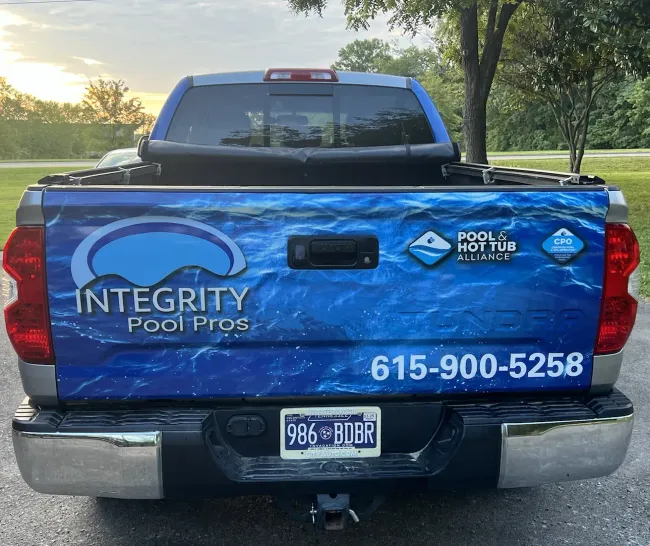 Blue pickup truck tailgate with Integrity Footprint and A Home logo, parked outdoors at dusk.