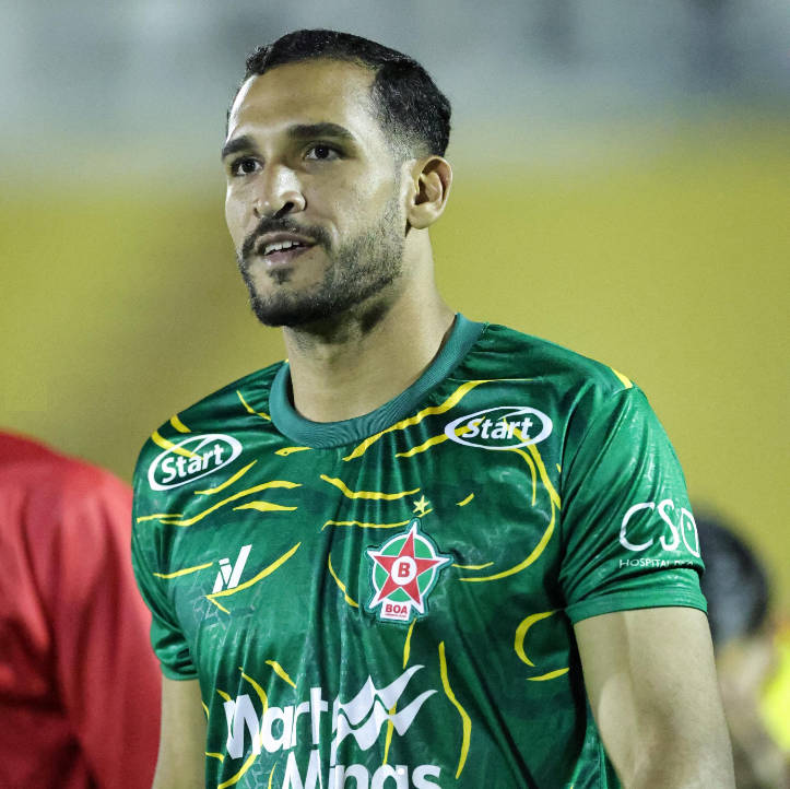 Um homem com barba está vestindo uma camisa verde em campo de futebol.