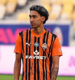 Homem com camisa preta e laranja olhando para o lado em um campo de futebol.