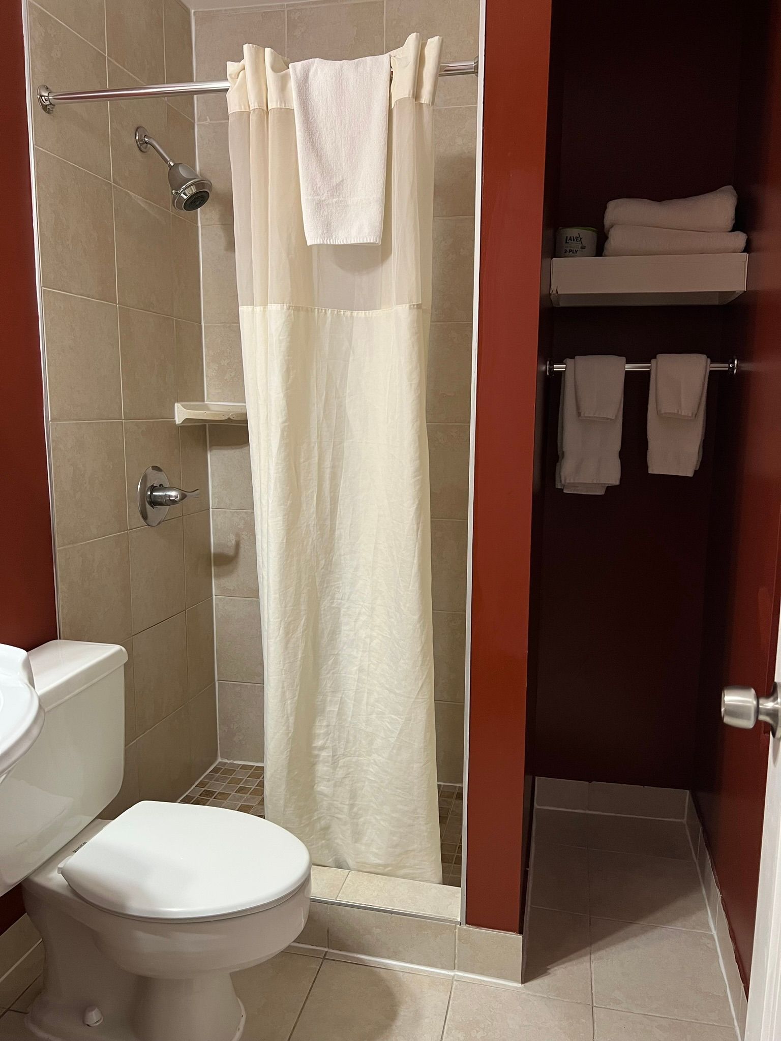 Small bathroom with a shower, toilet, and towel storage. Red walls and light tan tiles.