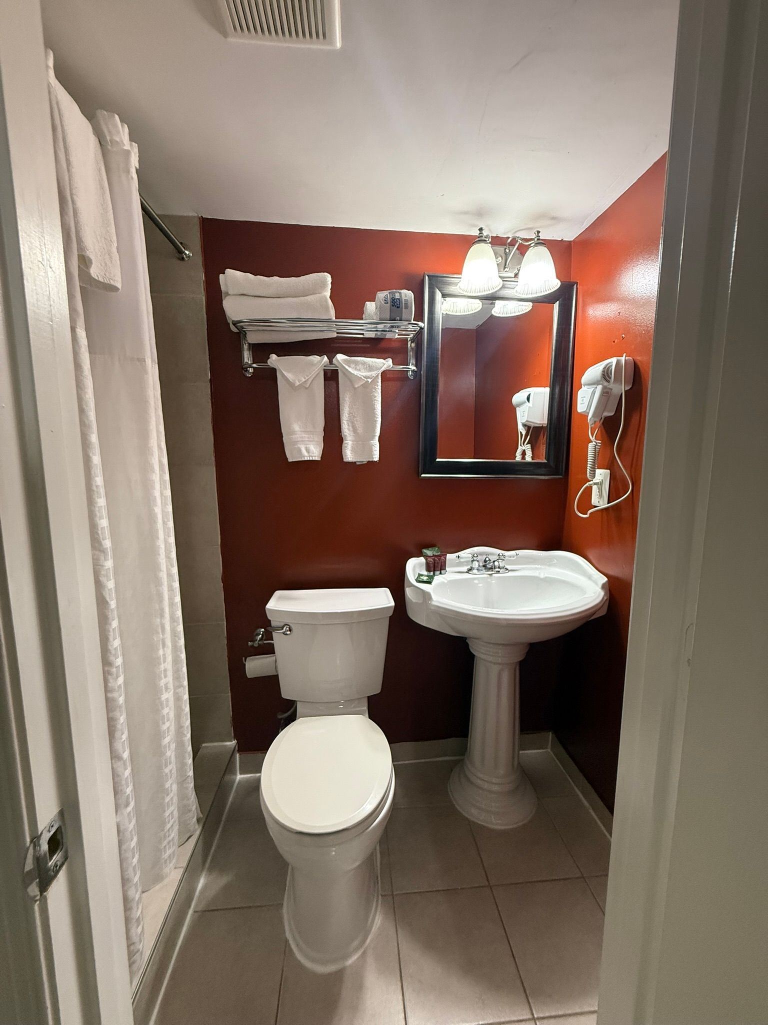 Small bathroom with a toilet, sink, and shower. Dark red wall behind the sink and a mirror. White towels are neatly hung.