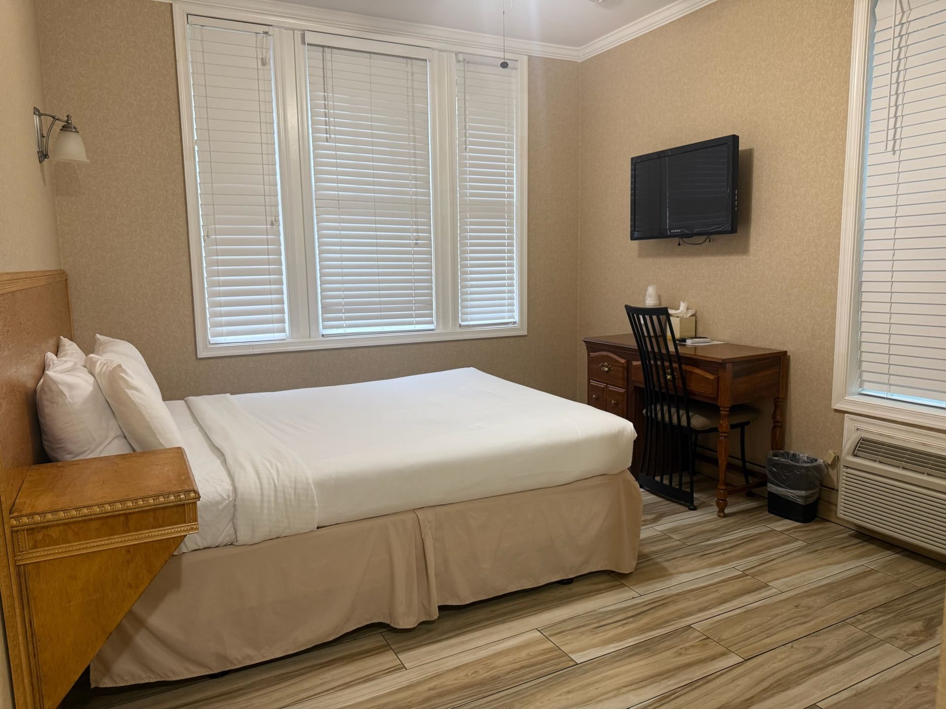 A hotel room featuring a bed with white linens, a desk with a TV, and a window with blinds.
