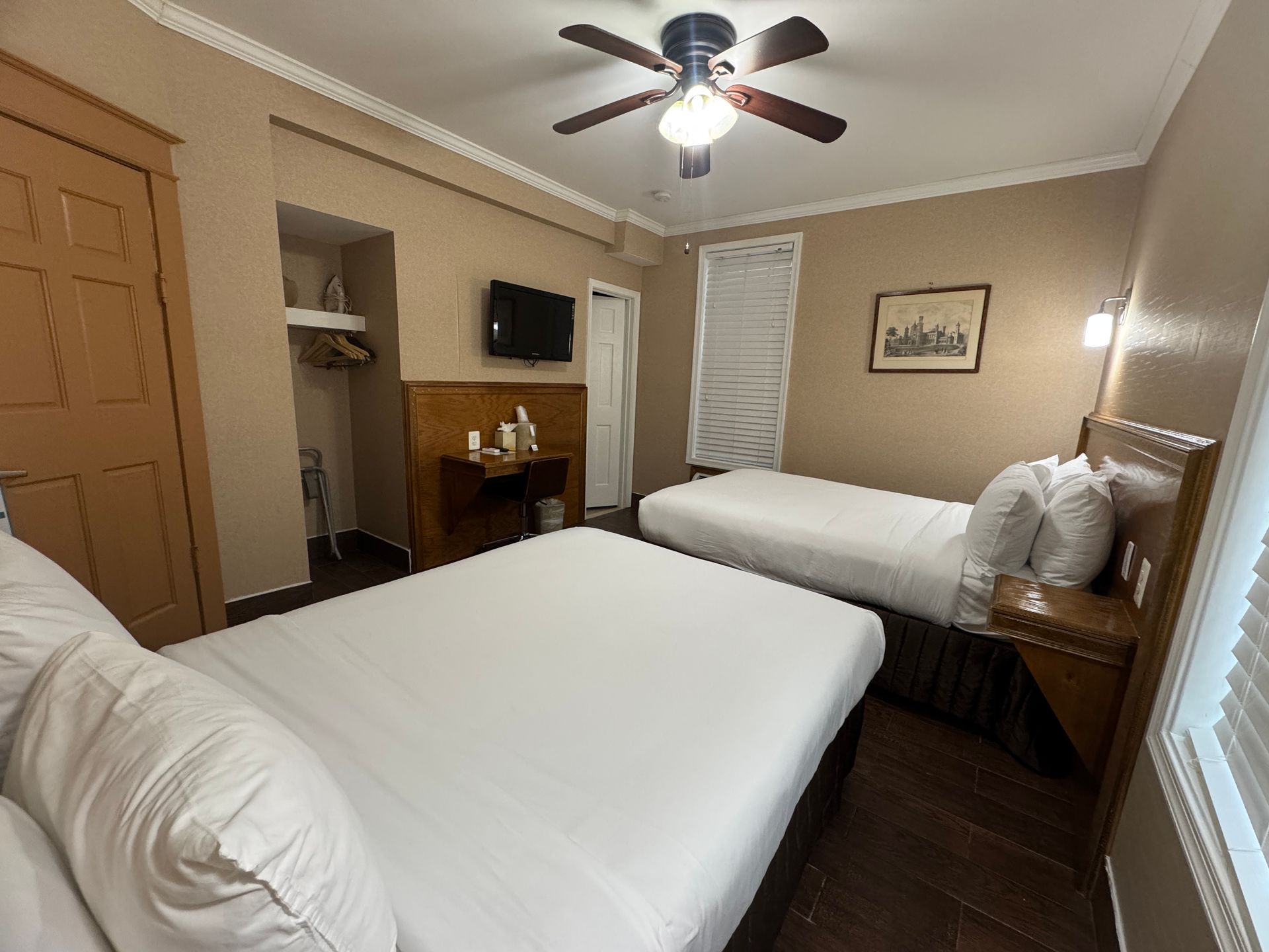 Hotel room with two beds, a small TV, and a ceiling fan. Wooden furniture and dark floors.