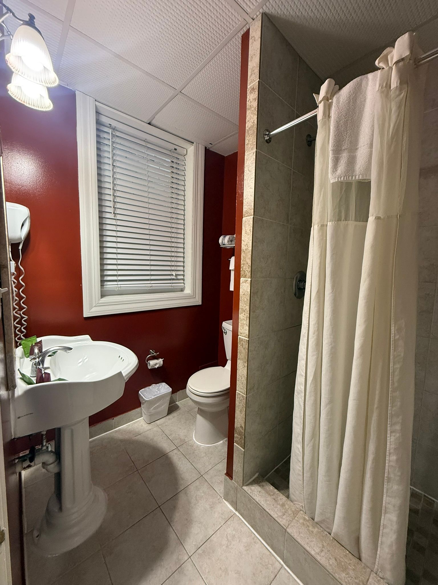 Bathroom with red walls, a white sink, toilet, and shower with a white curtain. A window with blinds is also present.