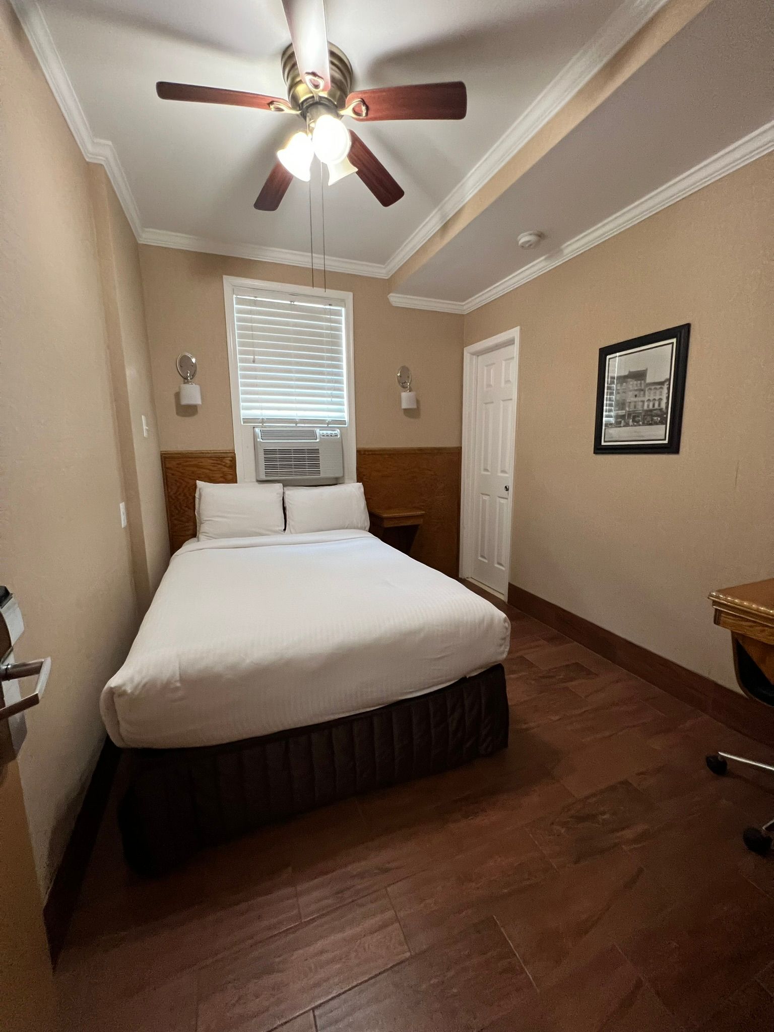 Small hotel room with a double bed, air conditioner, and a closed door. Brown wood floors and a framed picture on the wall.