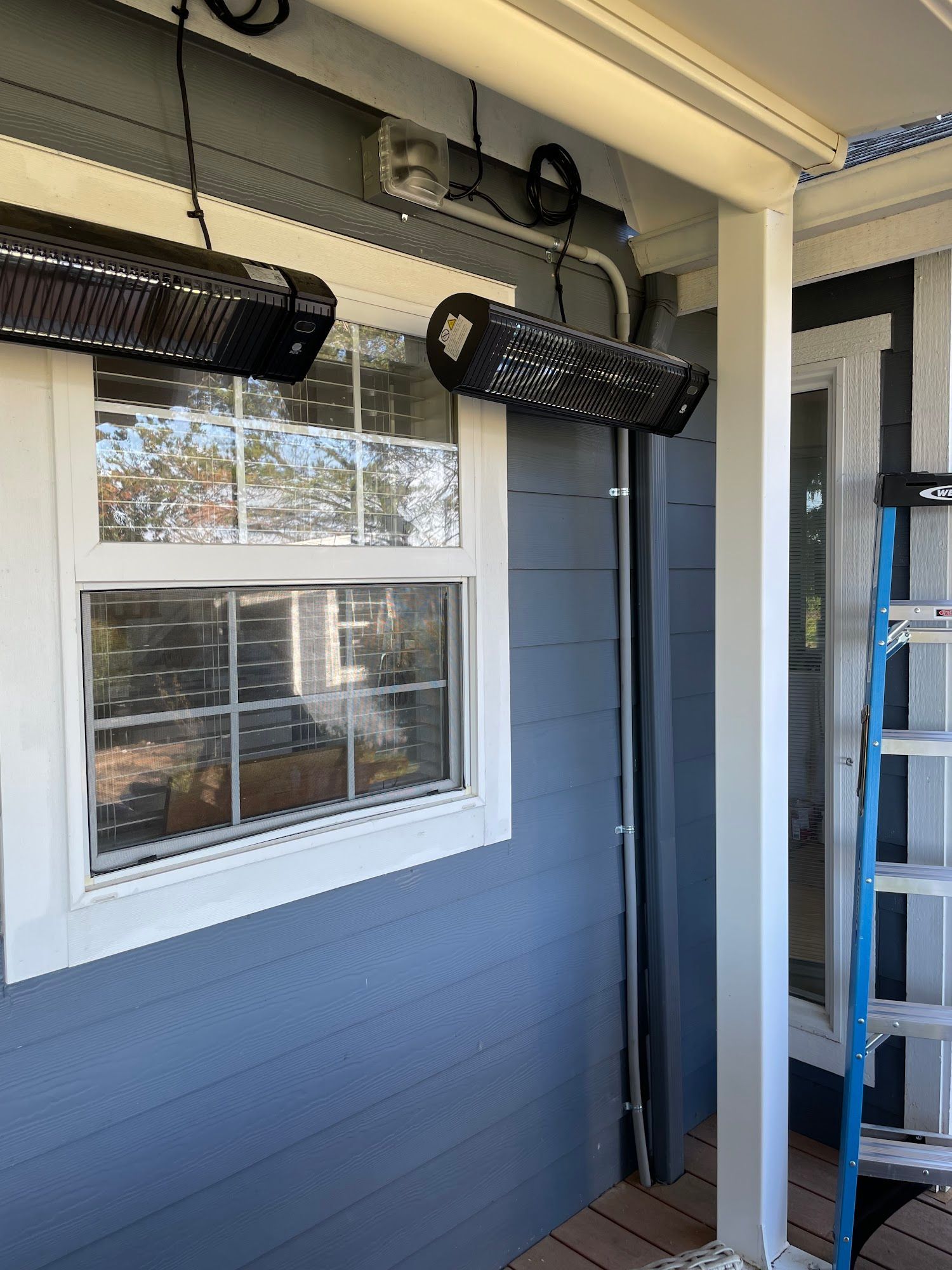 Two black outdoor heaters mounted on a porch, near a window and support column.