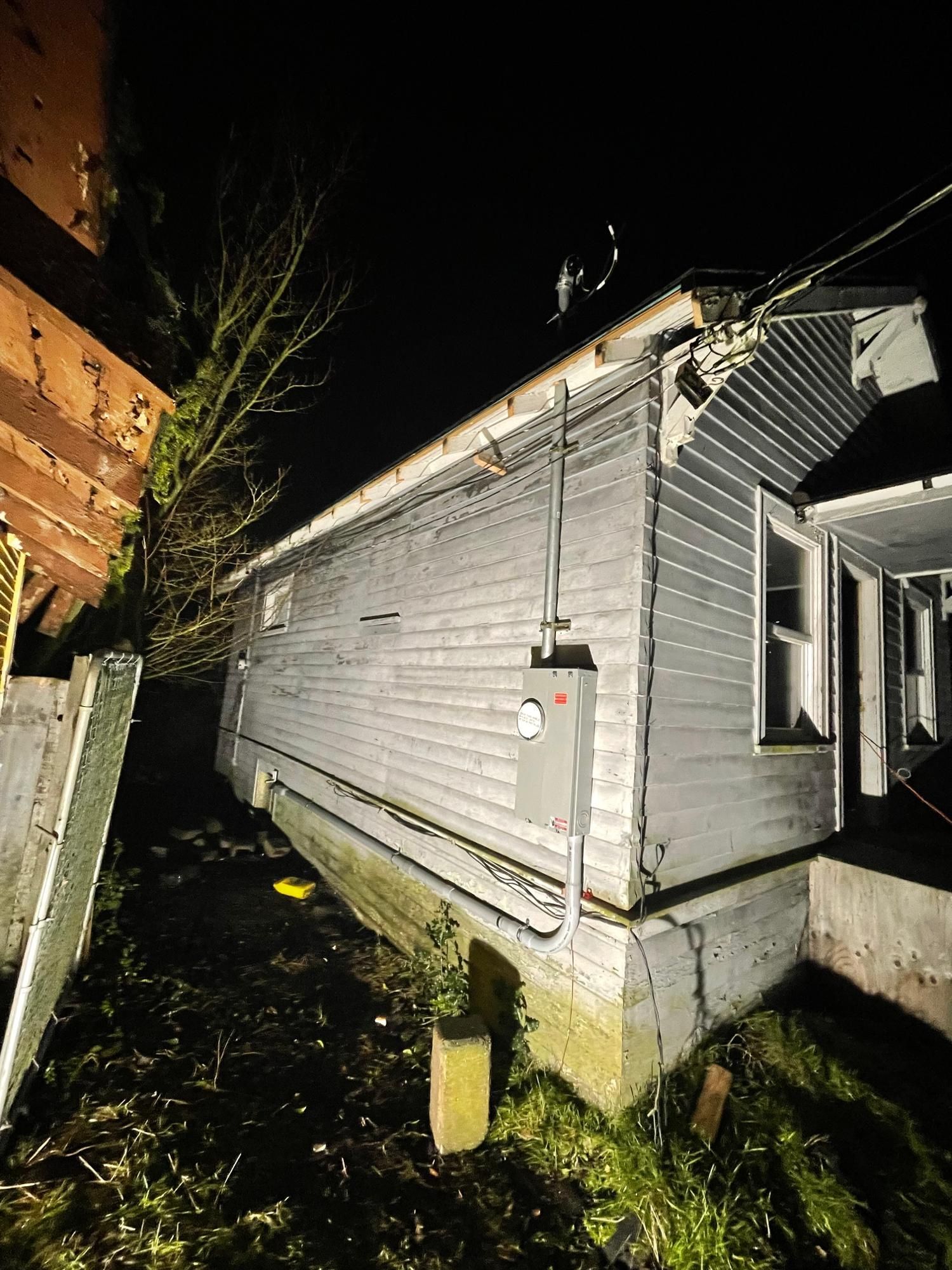 Exterior view of a weathered gray building at night. A breaker box and pipes are visible.