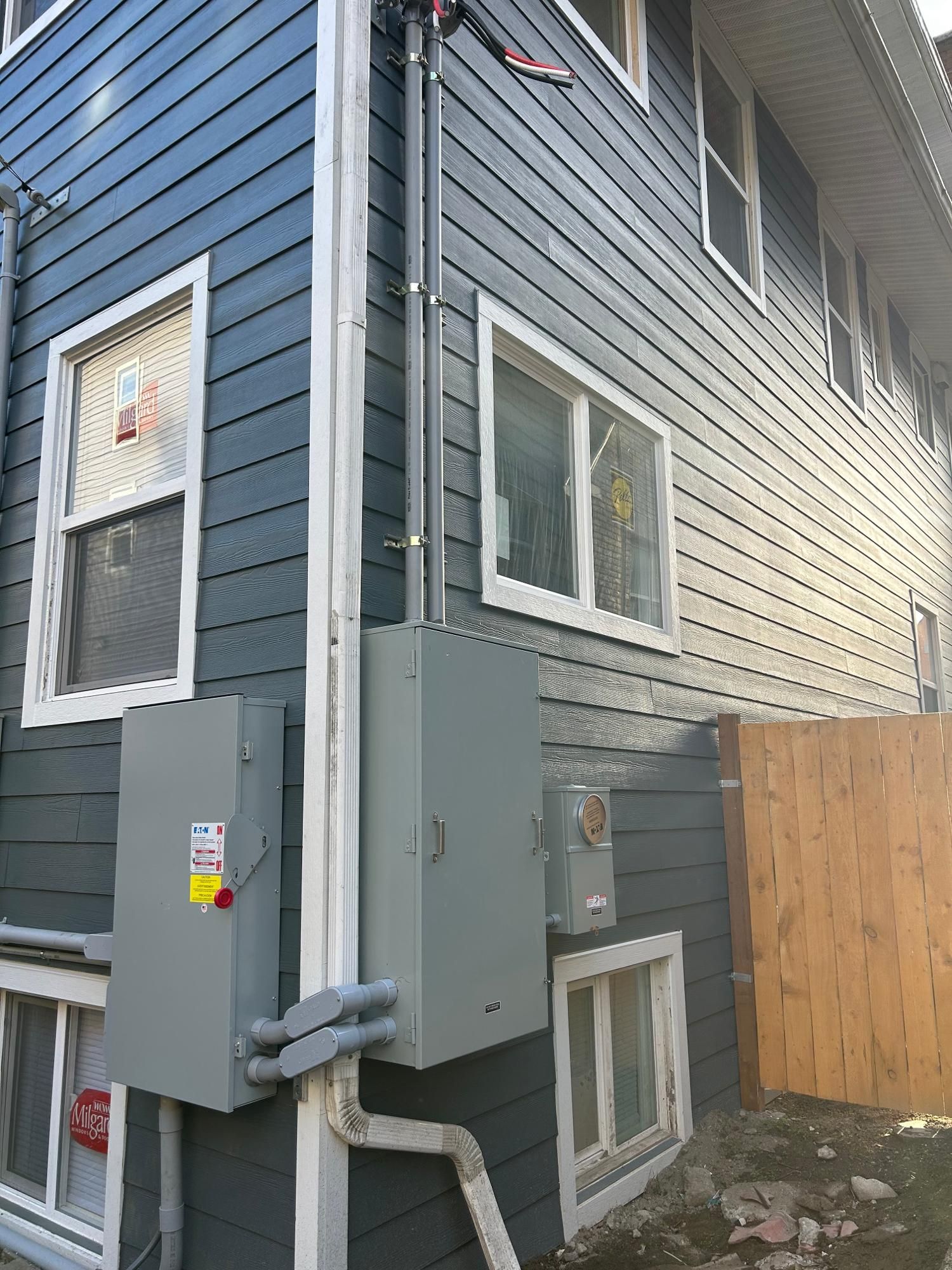 Exterior of a building with blue siding, gray electrical boxes, and conduits. White window frames and a wooden fence