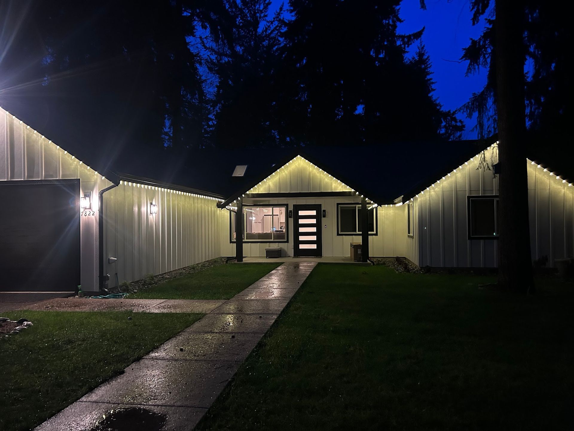 White house at night with string lights lining the roof and garage, a dark front door, and a walkway.